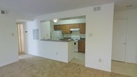 This single-floor space features an open and functional layout that connects the living area to the kitchen through a half-wall pass-through counter, creating a sense of openness while maintaining a defined separation between spaces. The living area is finished with wall-to-wall carpeting in a neutral beige tone, contributing to a warm and understated ambiance. Overhead vents are integrated into the ceiling, ensuring efficient climate control throughout.

The kitchen is efficiently arranged with warm-toned oak cabinetry lining both the upper and lower sections of the walls, providing ample storage. White appliances, including a refrigerator, electric range with oven, and a range hood above the cooktop, are standard features. The countertops are finished in a dark laminate, offering a subtle contrast to the lighter cabinetry. The kitchen floor transitions to a smooth, easy-to-clean surface, practical for the cooking area. A ceiling-mounted light fixture provides direct illumination over the kitchen workspace. The pass-through counter doubles as a convenient breakfast bar or serving surface, enhancing the utility of the shared living and kitchen space.