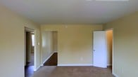 This single-floor residence offers a straightforward and functional layout designed for comfortable everyday living. The main living area is finished with wall-to-wall carpeting that adds a soft, warm feel underfoot, while the pale yellow walls lend the space a bright and welcoming ambiance. White baseboards and trim provide clean contrast against the neutral tones throughout.

An open doorway leads to a connecting hallway or adjoining room, where hardwood-style flooring is visible, creating a pleasant contrast in materials between spaces. Natural light filters in through a window visible in the adjacent area, contributing to an airy atmosphere. A second doorway with a standard white interior door offers access to another room, suggesting a well-connected and accessible floor plan.

The ceilings are smooth and of a standard height, keeping the space feeling open without being overwhelming. The overall design is clean and understated, providing a neutral canvas that is easy to personalize. The transition between the carpeted living space and the hard-surface hallway highlights the thoughtful separation of functional zones within the home. This space is well-suited for those seeking a simple, move-in-ready environment with a calm and neutral aesthetic.