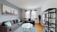 Located at 3804 Wabash Ave in Baltimore, MD, this single-floor residence offers a thoughtfully laid-out interior with warm hardwood flooring running throughout. The living room serves as the centerpiece of the home, featuring a calming gray and teal color palette, two gray tufted sofas accented with teal throw pillows, a round glass coffee table, and a decorative patterned area rug. A bold accent wall in slate blue anchors the room, complemented by large windows dressed in sheer curtains that allow generous natural light to fill the space. The primary bedroom is comfortably sized, featuring a window with an exterior view, a wall-mounted HVAC unit, and ample room for a full dresser and seating. A second bedroom offers a cozy, well-lit space with hardwood floors and a closet. The kitchen is efficiently designed with white cabinetry, dark countertops, a stainless steel sink, and a gas range, providing all the essentials for meal preparation. The full bathroom features classic white subway tile with dark accent trim, a window for natural light and ventilation, and a tub-shower combination. Overall, the space presents a clean, functional, and inviting atmosphere suited for comfortable everyday living.