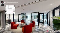 Located at 526 E Main St in the heart of Durham, NC, this thoughtfully designed office suite offers a compelling blend of modern aesthetics and functional workspace. The single-floor layout accommodates a generous footprint across three distinct areas, each flowing seamlessly into the next.

The central lounge serves as the social and visual heart of the space, anchored by bold red upholstered seating arranged around a low circular coffee table. Underfoot, a striking geometric patterned tile — featuring triangular shapes in grey, white, and charcoal — adds dramatic visual interest and defines the collaborative zone. A classic fluted column punctuates the open area, complementing the clean white ceiling with recessed and linear lighting.

Flanking the lounge are glass-walled private offices and a conference room enclosed by sleek black-framed floor-to-ceiling glass partitions, offering visual transparency while maintaining acoustic privacy. The conference room features a round table, ergonomic seating, abundant natural light, and large windows with views of the surrounding Durham cityscape.

A connecting corridor transitions to warm hardwood flooring, lined with curated large-format artwork and tall indoor plants in cylindrical planters, lending a gallery-like atmosphere. Elegant arched doorways add architectural character to the passageways. The overall ambiance is polished, contemporary, and sophisticated — well-suited for a professional and creative work environment.