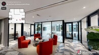 Located at 526 E Main St in the heart of Durham, NC, this thoughtfully designed office suite offers a compelling blend of modern aesthetics and functional workspace. The single-floor layout accommodates a generous footprint across three distinct areas, each flowing seamlessly into the next.

The central lounge serves as the social and visual heart of the space, anchored by bold red upholstered seating arranged around a low circular coffee table. Underfoot, a striking geometric patterned tile — featuring triangular shapes in grey, white, and charcoal — adds dramatic visual interest and defines the collaborative zone. A classic fluted column punctuates the open area, complementing the clean white ceiling with recessed and linear lighting.

Flanking the lounge are glass-walled private offices and a conference room enclosed by sleek black-framed floor-to-ceiling glass partitions, offering visual transparency while maintaining acoustic privacy. The conference room features a round table, ergonomic seating, abundant natural light, and large windows with views of the surrounding Durham cityscape.

A connecting corridor transitions to warm hardwood flooring, lined with curated large-format artwork and tall indoor plants in cylindrical planters, lending a gallery-like atmosphere. Elegant arched doorways add architectural character to the passageways. The overall ambiance is polished, contemporary, and sophisticated — well-suited for a professional and creative work environment.