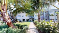 Located within a well-maintained multi-story building identified as 2500, this single-floor space is set amid a richly landscaped tropical environment. The building's exterior is finished in a clean white facade, complemented by balconies and large windows that allow for ample natural light throughout. The approach to the main entrance is framed by towering palm trees, manicured lawns, and an abundance of tropical foliage, creating a serene and inviting atmosphere before even stepping inside. A paved walkway guides visitors directly to the glass-door entry, which opens into the building's interior. The grounds reflect careful attention to landscaping, with a diverse mix of palms, broadleaf plants, and ground cover that contribute to the lush, resort-like character of the property. The single-floor layout offers the convenience of easy accessibility within the building. While interior imagery is limited due to lighting conditions in the available photos, the overall setting suggests a well-established property that balances architectural simplicity with natural tropical beauty. The combination of the bright white exterior, verdant surroundings, and thoughtfully maintained outdoor spaces positions this property as a distinctive and appealing space within its tropical locale.