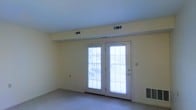 This single-floor space presents a clean and functional layout highlighted by a standout pair of French doors that serve as the room's focal point. The doors are fitted with horizontal blinds and feature multi-pane glass panels that invite generous amounts of natural light into the interior, creating a bright and open atmosphere. The doors appear to lead to an outdoor area, such as a patio or balcony, offering convenient access to the exterior.

The walls are finished in a soft, neutral cream tone that complements the light-colored carpet flooring, giving the space a cohesive and understated aesthetic. The ceiling is smooth and white, contributing to the overall sense of openness. Multiple HVAC vents are visible along the ceiling and near the baseboard, indicating a well-distributed climate control system throughout the space.

A wall-mounted heating unit is also present near the base of the French doors, ensuring warmth during cooler months. The room is free of interior clutter, making it easy to envision a variety of furniture arrangements. Overall, the space offers a comfortable, move-in-ready environment with practical features and a light, airy ambiance suited to everyday living.