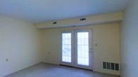 This single-floor space presents a clean and functional layout highlighted by a standout pair of French doors that serve as the room's focal point. The doors are fitted with horizontal blinds and feature multi-pane glass panels that invite generous amounts of natural light into the interior, creating a bright and open atmosphere. The doors appear to lead to an outdoor area, such as a patio or balcony, offering convenient access to the exterior.

The walls are finished in a soft, neutral cream tone that complements the light-colored carpet flooring, giving the space a cohesive and understated aesthetic. The ceiling is smooth and white, contributing to the overall sense of openness. Multiple HVAC vents are visible along the ceiling and near the baseboard, indicating a well-distributed climate control system throughout the space.

A wall-mounted heating unit is also present near the base of the French doors, ensuring warmth during cooler months. The room is free of interior clutter, making it easy to envision a variety of furniture arrangements. Overall, the space offers a comfortable, move-in-ready environment with practical features and a light, airy ambiance suited to everyday living.