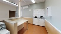 This shared laundry facility is a well-equipped, single-floor space designed for practical, everyday use. The room features terracotta-tiled flooring throughout, providing a durable and easy-to-clean surface that complements the neutral, two-tone wall treatment—cream on the upper half and a warm tan on the lower half—creating a clean, utilitarian aesthetic.

The facility is outfitted with multiple top-loading washing machines and stacked washer-dryer units, offering users ample options for completing laundry efficiently. Coin-operated controls are clearly visible on the machines, indicating a shared-use setup. Extended laminate folding counters run along one wall, providing generous workspace for sorting and folding laundry. A large wall mirror above the counter area enhances the sense of space within the room.

Overhead fluorescent lighting ensures the entire space is well-lit, while a sliding glass door along one wall invites natural light and provides ventilation. A floor drain is centrally located, adding a practical element for maintenance. The textured ceiling and visible ventilation panels complete the functional design of this straightforward, communal laundry space.