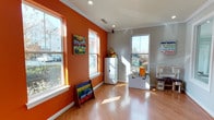 This single-floor space is defined by its welcoming and energetic character, centered around a thoughtfully designed playroom that immediately captures attention. The room features bold orange accent walls that create a warm, stimulating atmosphere, complemented by crisp white trim and crown molding that lend a polished, finished look throughout.

The flooring consists of warm-toned hardwood that runs seamlessly across the space, adding both durability and visual warmth. Multiple large windows with white frames flood the interior with generous natural light, casting bright, cheerful sunlight across the room while offering views of the surrounding neighborhood outside.

The layout is open and spacious, providing ample room for movement and activity. White storage furniture is neatly arranged to keep the space organized, while a colorful book display rack adds to the room's playful aesthetic. Decorative wall art with vibrant, child-friendly themes reinforces the room's fun and inviting character.

Recessed ceiling lighting supplements the abundant natural light, ensuring the space remains well-lit at all times. The combination of the vivid color palette, quality finishes, and thoughtful layout makes this a functional and engaging environment designed to inspire creativity and play.