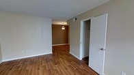 This single-story space is characterized by a clean, functional layout with warm hardwood-style flooring running throughout. The main living area features neutral beige walls and a textured popcorn ceiling, creating a simple and understated backdrop. An open passageway connects the living space to an adjacent dining area, which is distinguished by a rich, darker brown accent wall and a pendant light fixture that adds a focal point to the room. The transition between spaces flows naturally, contributing to an open and connected feel. A closet with a built-in rod is conveniently situated off the main living area, offering practical storage. A visible HVAC vent in the ceiling indicates climate control throughout the space. The overall ambiance is modest and move-in ready, with cohesive warm tones tying the rooms together.