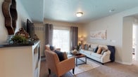 This single-floor residence offers a thoughtfully designed living space that balances comfort and style. The living room serves as the heart of the home, showcasing a neutral color palette that creates a warm and inviting atmosphere. A large sliding glass door framed by elegant grommet-top drapes floods the space with natural light, offering a bright and airy feel. The textured ceiling adds a subtle architectural detail overhead, complemented by flush-mount lighting. The carpeted floor provides a soft underfoot experience, anchored by a plush area rug that defines the seating area. The living space is arranged with a cushioned sofa and coordinating accent chairs in warm tones, centered around a sleek rectangular coffee table. Decorative throw pillows in navy, rust, and neutral tones add tasteful pops of color. A media console along one wall provides functional storage and display space. Abstract wall art contributes a contemporary touch to the overall décor. The layout flows naturally, making the space feel open and well-proportioned. The overall ambiance is casual yet polished, making it a comfortable and functional everyday living environment.