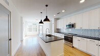 This single-floor residence showcases a clean, contemporary design with a cohesive color palette of white and dark accents carried throughout the space. The heart of the home is an open-concept kitchen that flows seamlessly into the living area, allowing natural light to pour in through large front-facing windows. The kitchen is well-equipped with stainless steel appliances, crisp white shaker-style cabinetry, dark countertops, and a striking herringbone tile backsplash. A generously sized center island with a dark countertop and an integrated sink serves as both a functional workspace and a focal point, illuminated by a pair of bold matte black pendant lights overhead. Warm-toned hardwood-style flooring extends throughout the main living areas, adding a sense of continuity and warmth. The bedrooms are carpeted for comfort, each benefiting from natural light and ceiling fans. The primary bedroom is complemented by a spacious walk-in closet outfitted with multi-tier wire shelving. Both bathrooms echo the kitchen's design language, featuring white vanities topped with dark countertops, large framed mirrors, and a clean tile surround in the tub and shower area. The overall ambiance is bright, modern, and move-in ready.