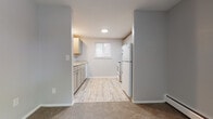 This single-floor residential unit offers a straightforward and functional layout across several well-defined areas. The living spaces are finished with wall-to-wall neutral beige carpeting, complemented by light gray walls and white baseboards throughout, creating a clean and cohesive aesthetic. Baseboard heating units are present in multiple rooms, ensuring efficient climate control, while a wall-mounted air conditioning unit in the main living area provides additional temperature management. The living room receives natural light through large windows with horizontal blinds and features a ceiling fan with integrated lighting. A second carpeted room offers two windows on adjacent walls, flooding the space with natural light from multiple directions. The kitchen is a distinct area with light wood-look vinyl flooring, gray lower cabinetry, upper cabinets, a white refrigerator, a white range, and a window that brings in additional natural light. The bathroom is tiled in classic white subway-style tile from floor to mid-wall, and includes a white vanity with under-cabinet storage, a mirrored medicine cabinet, and chrome fixtures. An entry area features a warm wood-toned door alongside a spacious built-in closet with bifold doors, providing ample storage at the entry point of the unit.