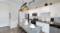 Highland Station presents a well-crafted single-floor residence that seamlessly blends modern finishes with comfortable living. The kitchen serves as a standout feature, showcasing crisp white shaker cabinetry extending to the ceiling, dark granite countertops, a classic white subway tile backsplash, and a full suite of stainless steel appliances including a range, microwave, dishwasher, and refrigerator. A generously sized kitchen island with matching granite provides additional prep space and casual seating, while stylish pendant lighting adds a contemporary touch overhead. Track lighting further illuminates the workspace throughout. The living area offers a warm and inviting atmosphere centered around a sleek built-in electric fireplace framed by a white mantel. Large windows allow natural light to fill the room, complemented by wood-look flooring that flows throughout the main areas. The bedroom is a peaceful retreat featuring plush carpeting, generous window views, and a ceiling fan. The primary bathroom connects directly to a walk-in closet, and features a white vanity with dark countertops and a well-lit mirror. The walk-in closet provides ample storage with dedicated hanging space. Throughout the residence, neutral tones, quality finishes, and thoughtful design details create a cohesive and polished living environment.