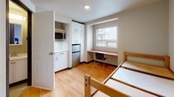 Located at 4743 21st Ave NE in Seattle's University District, this compact studio unit is thoughtfully designed to maximize every square foot of available space. The main living area features warm light hardwood flooring that runs throughout, creating a cohesive and inviting atmosphere. A built-in kitchenette is neatly integrated along one wall, equipped with white cabinetry extending to the ceiling for ample storage, a stainless steel compact refrigerator, a microwave, and a small countertop workspace — providing all essential cooking conveniences in a streamlined layout. Adjacent to the kitchenette, a recessed built-in desk nook frames a window that allows natural light to filter in, offering an ideal dedicated study or work area. The sleeping area accommodates a bed frame and benefits from a wall-mounted HVAC unit, ensuring year-round comfort. The private bathroom is clean and functional, featuring a toilet with overhead storage cabinetry, a vanity with sink, a shower stall, and chrome fixtures. A closet provides additional storage solutions. The overall design is clean and minimal, with neutral tones, white walls, and simple finishes that give the space a bright, orderly feel well-suited for a focused, efficient lifestyle.