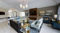 This single-story space showcases a thoughtfully designed living area that blends contemporary elegance with refined comfort. The room is anchored by a striking entertainment wall featuring a warm walnut-toned wood panel that frames a wall-mounted flat-screen television, flanked by a coordinating media console and complementary landscape artwork. Textured wallcovering in a subtle herringbone pattern adds depth to the walls, contributing to the room's layered aesthetic.

The seating arrangement is both stylish and functional, combining a plush charcoal sofa with shimmering teal and silver accent chairs and a deep navy accent chair. Decorative throw pillows in coordinating tones tie the palette together seamlessly. A gold and glass coffee table serves as a centerpiece, adorned with curated decorative accessories including a starburst sculpture and metallic accents.

Overhead, a cluster of cylindrical pendant lights with gold hardware provides warm, ambient illumination, complemented by recessed ceiling lighting. Light-toned tile flooring anchors a richly patterned area rug beneath the seating area. Natural light from French doors at the far end brightens the space, enhancing its open and welcoming ambiance.