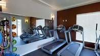 This single-floor fitness center is thoughtfully designed to provide a functional and motivating workout environment. The space is anchored by a row of modern treadmills and cardio machines, offering users a variety of options for cardiovascular exercise. A full-length mirrored wall runs along one side of the room, effectively doubling the perceived sense of space while allowing users to monitor their form during workouts. The bold, deep red-brown accent wall adds a dynamic visual contrast against the neutral white walls and ceiling, creating an energizing atmosphere. Recessed tray ceiling detailing adds a touch of architectural refinement to the space. Wall-mounted sconce lighting provides a warm, focused glow throughout the room, complementing the natural light that filters in through the window treatments. The flooring is finished in a dark, durable fitness-grade carpet, well-suited for high-traffic use. Overall, the fitness center strikes a balance between practicality and aesthetic appeal, making it a welcoming space for daily exercise routines.