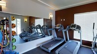 This single-floor fitness center is thoughtfully designed to provide a functional and motivating workout environment. The space is anchored by a row of modern treadmills and cardio machines, offering users a variety of options for cardiovascular exercise. A full-length mirrored wall runs along one side of the room, effectively doubling the perceived sense of space while allowing users to monitor their form during workouts. The bold, deep red-brown accent wall adds a dynamic visual contrast against the neutral white walls and ceiling, creating an energizing atmosphere. Recessed tray ceiling detailing adds a touch of architectural refinement to the space. Wall-mounted sconce lighting provides a warm, focused glow throughout the room, complementing the natural light that filters in through the window treatments. The flooring is finished in a dark, durable fitness-grade carpet, well-suited for high-traffic use. Overall, the fitness center strikes a balance between practicality and aesthetic appeal, making it a welcoming space for daily exercise routines.