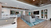 Located at 3680 46th Ave S in Saint Petersburg, Florida, this single-floor space is a beautifully designed coastal-inspired lounge and entertainment venue. The open floor plan is anchored by wood-look tile flooring that flows seamlessly throughout, tying together the various functional areas with a warm, cohesive aesthetic.

The centerpiece of the space is a generously sized media and lounge area outfitted with rows of premium leather recliner-style seating arranged theater-fashion, all oriented toward a large wall-mounted flat-screen TV set within a custom built-in shelving unit. The built-ins provide stylish display space and add architectural depth to the room.

Overhead, a striking tray ceiling features rich wood-paneled insets complemented by sleek ceiling fans and recessed lighting, lending the space a refined yet relaxed ambiance. Large sliding glass doors and windows along the perimeter flood the interior with abundant natural light and offer views of the surrounding Florida landscape, including swaying palms.

A well-appointed bar and kitchenette area serves as a social hub, complete with crisp white cabinetry, a quartz countertop with bar seating, and a vibrant ocean-blue mosaic tile backsplash. Floating wood shelves and teal pendant lighting reinforce the coastal theme throughout. A foosball table and additional seating areas round out the space, making it ideal for gatherings and community entertainment.