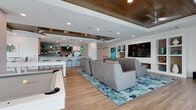 Located at 3680 46th Ave S in Saint Petersburg, Florida, this single-floor space is a beautifully designed coastal-inspired lounge and entertainment venue. The open floor plan is anchored by wood-look tile flooring that flows seamlessly throughout, tying together the various functional areas with a warm, cohesive aesthetic.

The centerpiece of the space is a generously sized media and lounge area outfitted with rows of premium leather recliner-style seating arranged theater-fashion, all oriented toward a large wall-mounted flat-screen TV set within a custom built-in shelving unit. The built-ins provide stylish display space and add architectural depth to the room.

Overhead, a striking tray ceiling features rich wood-paneled insets complemented by sleek ceiling fans and recessed lighting, lending the space a refined yet relaxed ambiance. Large sliding glass doors and windows along the perimeter flood the interior with abundant natural light and offer views of the surrounding Florida landscape, including swaying palms.

A well-appointed bar and kitchenette area serves as a social hub, complete with crisp white cabinetry, a quartz countertop with bar seating, and a vibrant ocean-blue mosaic tile backsplash. Floating wood shelves and teal pendant lighting reinforce the coastal theme throughout. A foosball table and additional seating areas round out the space, making it ideal for gatherings and community entertainment.