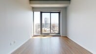 This contemporary high-rise unit is thoughtfully designed across a single floor, offering a clean, modern aesthetic throughout. Light hardwood flooring flows through the main living areas, complemented by neutral gray walls that create a bright and airy atmosphere. The open-plan layout connects the living space to a well-appointed kitchen featuring rich dark wood cabinetry extending to the ceiling, under-cabinet lighting, a white countertop with a breakfast bar overhang, and stainless steel fixtures — a functional and stylish centerpiece of the unit. The bedroom areas are generously sized, with one room finished in soft carpeting and the other in hardwood flooring. Both feature expansive floor-to-ceiling sliding windows that frame impressive panoramic city skyline views, flooding the rooms with abundant natural light. The unit includes two bathrooms, each featuring white subway-tiled tub-and-shower combinations, light stone-look tile flooring, and dark wood vanity cabinetry with chrome hardware. Additional storage is provided by a built-in closet with louvered bifold doors near the entryway. The overall ambiance is clean, contemporary, and urban, with a neutral palette and high-quality finishes that offer a refined city living experience.