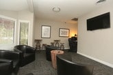 This single-floor commercial space in Macon, Georgia, is designed to offer a comfortable and functional environment for both relaxation and productivity. The main lounge area is finished with patterned carpet tiles in a neutral gray tone, complemented by clean white baseboards and warm taupe walls that create a professional yet inviting atmosphere. Large windows fitted with white horizontal blinds allow natural light to fill the room, offering views of the surrounding exterior. A wall-mounted flat-screen television serves as a central focal point, making the space well-suited for presentations or casual viewing. Seating is arranged thoughtfully throughout, with a cluster of dark leather barrel chairs grouped around a rustic wooden side table, alongside high-top tables with industrial-style stools that provide additional flexible seating options. Framed Macon, Georgia-themed artwork adorns the walls, lending a distinct local character to the space. A dedicated workspace tucked off the main lounge features a desk and ergonomic chair, ideal for individual work or administrative tasks. The entry area transitions from tile flooring to carpet and features a glass-paneled door that enhances the flow of natural light. Overall, the space strikes a balance between a welcoming lounge and a practical office environment.