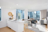 This thoughtfully designed single-floor residence offers a seamless blend of modern aesthetics and urban living. The open-concept layout connects the living, dining, and kitchen areas, all bathed in abundant natural light streaming through expansive floor-to-ceiling windows that frame striking city skyline views. Light hardwood flooring runs throughout, enhancing the bright, airy ambiance.

The contemporary kitchen is outfitted with high-gloss white cabinetry, stainless steel appliances, a large central island with a built-in sink, and additional windows that capture sweeping cityscape panoramas. The dining area comfortably accommodates multiple guests and flows naturally into the living room, which features oversized glazing that fills the space with sunlight and unobstructed views of the surrounding urban skyline.

The residence includes multiple bedrooms, each with large windows showcasing city views. One bedroom features warm textured wall paneling and neutral tones, while another offers a clean, versatile layout suitable as a home office or den. A bright hallway connects the rooms, accented with curated artwork.

Two well-appointed bathrooms complete the residence. The primary bathroom features a dual-sink vanity with ample storage and a shower-tub combination, while the secondary bathroom includes a clean white vanity and tiled shower, maintaining the consistent modern aesthetic found throughout the home.