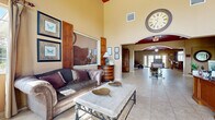 Located at 132 Carolina Lake Dr in Daytona Beach, FL, this single-story community clubhouse spans a generous footprint designed to accommodate a variety of activities and gatherings. The main lounge area immediately impresses with soaring ceilings, warm beige walls, and large tile flooring that extends throughout the space. A dramatic oversized wall clock serves as a focal point, complementing the traditionally styled furnishings, including a striped sofa, accent chairs, and an elegant marble-topped coffee table. Natural light filters in through multiple windows dressed with neutral drapes, offering pleasant outdoor views of the surrounding landscape.

The space transitions seamlessly into a media room featuring a bold burgundy ceiling, French doors leading outside, and a built-in entertainment console. Adjacent to this is a dining area with a classic wooden table and ornate chairs, positioned beneath recessed lighting. The kitchen is well-appointed with rich cherry wood cabinetry, light countertops, and a breakfast bar with upholstered bar seating. A dedicated computer room provides built-in workstations with carpeted flooring and natural light. Together, these areas create a functional, welcoming environment with a cohesive warm and traditional aesthetic throughout.