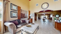 Located at 132 Carolina Lake Dr in Daytona Beach, FL, this single-story community clubhouse spans a generous footprint designed to accommodate a variety of activities and gatherings. The main lounge area immediately impresses with soaring ceilings, warm beige walls, and large tile flooring that extends throughout the space. A dramatic oversized wall clock serves as a focal point, complementing the traditionally styled furnishings, including a striped sofa, accent chairs, and an elegant marble-topped coffee table. Natural light filters in through multiple windows dressed with neutral drapes, offering pleasant outdoor views of the surrounding landscape.

The space transitions seamlessly into a media room featuring a bold burgundy ceiling, French doors leading outside, and a built-in entertainment console. Adjacent to this is a dining area with a classic wooden table and ornate chairs, positioned beneath recessed lighting. The kitchen is well-appointed with rich cherry wood cabinetry, light countertops, and a breakfast bar with upholstered bar seating. A dedicated computer room provides built-in workstations with carpeted flooring and natural light. Together, these areas create a functional, welcoming environment with a cohesive warm and traditional aesthetic throughout.