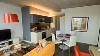 This single-floor space showcases a thoughtfully designed open-concept layout that seamlessly integrates the kitchen, dining, and living areas into one cohesive environment. The kitchen is fitted with dark grey cabinetry, stainless steel appliances including a built-in oven and microwave, and a dark granite countertop that runs the length of the galley-style cooking space. Warm under-cabinet and cove lighting casts an inviting amber glow across the kitchen, contrasting beautifully with the cool grey walls and exposed concrete ceiling above. The dining area features a round pedestal table surrounded by sleek white chairs, creating a clean and modern gathering spot. A framed urban artwork on the adjacent white wall adds a touch of city character to the space. The living area anchors the room with a bold mustard-yellow sofa adorned with patterned throw pillows, set atop a colorful area rug that introduces warmth and texture to the polished dark concrete flooring. The interplay of industrial elements — raw concrete, dark tones — with vibrant furnishings and warm lighting gives this space a dynamic, contemporary ambiance that feels both stylish and livable.