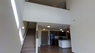 This two-story residence makes a striking impression with its soaring double-height ceilings that flood the main living area with a sense of openness and grandeur. The main floor is designed with an open-concept layout, seamlessly connecting the living area to the kitchen. The kitchen is well-equipped, featuring dark cabinetry, a black refrigerator, and a peninsula with a sink and countertop seating, all illuminated by a series of pendant lights that add a modern, stylish touch. Flush-mounted ceiling lights provide additional ambient lighting throughout the space. Rich, dark hardwood-style flooring spans the main level, offering a warm contrast to the crisp white walls. A carpeted staircase along one wall leads to the upper level, where a loft-style space overlooks the main floor below via an open railing. This elevated vantage point enhances the dramatic vertical scale of the interior. The overall ambiance is clean, contemporary, and functional, with a neutral color palette that creates a versatile backdrop suitable for a variety of interior styles. The thoughtful layout and architectural details make this a distinctive and inviting two-floor living space.