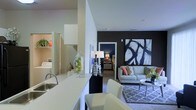 This single-floor residence showcases a thoughtfully designed open-concept layout that integrates the kitchen and living areas into one cohesive space. The kitchen features a clean, white countertop peninsula with an undermount sink and bar seating, creating a natural transition into the living area. White cabinetry and bright surfaces contribute to a light, airy atmosphere throughout.

The living room is anchored by a bold charcoal accent wall, serving as a dramatic backdrop for a large-scale abstract artwork in black and white. A light gray sofa dressed with colorful accent pillows sits atop a geometric patterned area rug, adding texture and visual interest to the space. A sleek round coffee table completes the seating arrangement.

A glimpse through a doorway reveals a bedroom with its own distinct, moodier color palette, suggesting well-defined private spaces within the home. To the side, a partially visible laundry area with stacked or side-by-side units indicates in-unit laundry convenience.

Overall, the space balances contemporary design with functional living, combining contrasting tones, curated décor, and practical amenities in a modern, well-appointed layout.