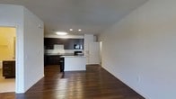 This single-floor space showcases a clean, contemporary design centered around an open-concept layout that seamlessly connects the living area to the kitchen. Rich, dark hardwood flooring runs throughout, providing a striking contrast against the crisp white walls and creating a warm yet sophisticated ambiance.

The kitchen is a standout feature, outfitted with dark espresso cabinetry, light stone countertops, and a full suite of black stainless steel appliances, including a refrigerator, range, and over-the-range microwave. A peninsula-style island extends from the kitchen, offering additional counter space and serving as a natural dividing point between the cooking and living areas.

Recessed lighting and a flush ceiling fixture illuminate the kitchen efficiently, while the open living area benefits from the overall brightness of the space. The neutral white walls and smooth ceilings provide a versatile backdrop, ideal for a range of interior styles. A doorway visible from the main area hints at additional rooms or storage beyond. The overall layout is practical and well-organized, making efficient use of the available footprint while maintaining an open, airy feel throughout.