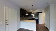 This single-floor residence showcases a thoughtfully designed open-concept layout that seamlessly blends style and functionality. The kitchen serves as the centerpiece of the space, featuring rich dark cabinetry that extends to the ceiling, creating a bold and contemporary aesthetic. Granite countertops with warm, speckled tones provide ample prep space and extend into a peninsula that offers additional seating and a natural division between the kitchen and living areas. A mosaic tile backsplash adds texture and visual interest, complementing the dark cabinetry and light countertops. Two industrial-style pendant lights hang above the peninsula, casting focused illumination and reinforcing the modern design language. Stainless steel appliances, including a gas range, built-in microwave, and refrigerator, complete the kitchen's polished look. The flooring throughout is a rich, dark hardwood that anchors the space and enhances its warm, contemporary character. Track lighting in the kitchen ensures the workspace is well-lit, while the overall open layout promotes a sense of spaciousness and easy flow. White interior doors and clean wall finishes provide a crisp contrast to the darker design elements, keeping the space feeling bright and balanced.