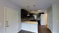 This single-floor residence showcases a thoughtfully designed open-concept layout that seamlessly blends style and functionality. The kitchen serves as the centerpiece of the space, featuring rich dark cabinetry that extends to the ceiling, creating a bold and contemporary aesthetic. Granite countertops with warm, speckled tones provide ample prep space and extend into a peninsula that offers additional seating and a natural division between the kitchen and living areas. A mosaic tile backsplash adds texture and visual interest, complementing the dark cabinetry and light countertops. Two industrial-style pendant lights hang above the peninsula, casting focused illumination and reinforcing the modern design language. Stainless steel appliances, including a gas range, built-in microwave, and refrigerator, complete the kitchen's polished look. The flooring throughout is a rich, dark hardwood that anchors the space and enhances its warm, contemporary character. Track lighting in the kitchen ensures the workspace is well-lit, while the overall open layout promotes a sense of spaciousness and easy flow. White interior doors and clean wall finishes provide a crisp contrast to the darker design elements, keeping the space feeling bright and balanced.