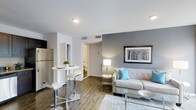 This single-floor unit at Deer Creek offers an open-concept layout that seamlessly connects the living and dining areas, creating a cohesive and functional living space. The interior is finished with wood-look flooring throughout, contributing warmth and a modern aesthetic that ties the rooms together.

The living area is highlighted by a neutral toned accent wall in grey, providing a stylish backdrop for the space. Recessed lighting and a table lamp offer layered illumination, while a framed nature-inspired artwork adds a decorative focal point. The living area comfortably accommodates a tufted sofa with coordinating accent pillows in soft blue and grey tones, alongside a glass-top coffee table.

Adjacent to the living area, a compact dining space features a tall bistro-style table with bar stools, ideal for casual dining. A stainless steel refrigerator is visible nearby, hinting at a functional kitchen. White walls and white trim throughout the unit maintain a clean, bright atmosphere. A hallway visible in the background leads to additional areas of the unit, including what appears to be a bathroom, rounding out this thoughtfully designed space.