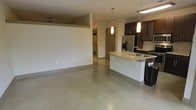 This single-floor space showcases a clean, contemporary design centered around an open-concept floor plan that seamlessly connects the kitchen, dining, and living areas. Polished concrete floors run throughout, lending an urban, industrial character while remaining easy to maintain.

The kitchen is a standout feature, equipped with dark espresso cabinetry, a stainless steel refrigerator, and a gas range. A curved granite-topped island takes center stage, offering a double stainless steel sink and pendant lighting above — making it both a functional workspace and a natural gathering point.

The living and dining areas are bathed in natural light, courtesy of large, multi-pane windows that frame outdoor views of surrounding greenery. Pendant and overhead lighting fixtures complement the natural illumination throughout the day. A ceiling fan is also present in the living area, adding to comfort and air circulation.

A hallway visible from the main living space leads to additional rooms, including a bathroom with modern fixtures. The layout is practical and efficient, with clean sightlines that make the interior feel open and airy. Overall, this space combines modern finishes, thoughtful design, and a bright, welcoming ambiance ideal for contemporary living.