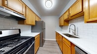 This single-floor residence offers a well-organized layout with distinct living, sleeping, dining, and kitchen zones connected by a straightforward open corridor. The galley-style kitchen is a functional highlight, featuring warm honey-toned wood cabinetry lining both walls, white subway tile backsplash, white countertops, stainless steel appliances including a gas range and full-size refrigerator, and a deep single-basin sink with a modern pull-down faucet. Wood-look luxury vinyl plank flooring runs throughout the main living and kitchen areas, lending a cohesive, easy-to-maintain aesthetic. The living area is generously proportioned and receives abundant natural light through a large floor-to-ceiling sliding glass door fitted with vertical blinds. The space connects seamlessly to additional living or dining areas, maintaining an open flow throughout. Bedrooms are carpeted in neutral gray tones, each equipped with windows dressed in blinds that allow for natural light and privacy. The bathroom features a full tub-shower combination with white tile surround, wood-look tile flooring, and clean white fixtures throughout. Neutral gray wall tones and white trim are consistent throughout the unit, creating a calm, cohesive interior palette.