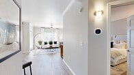 This single-story residence is designed with a clean, contemporary aesthetic throughout, characterized by light neutral tones, wood-look flooring, and textured ceilings with crown molding details. A welcoming entryway leads into the main living area, where a ceiling fan, large windows with bright white curtains, and a tiled fireplace create an inviting focal point. The space receives generous natural light, contributing to the open and airy feel of the room.

Adjacent to the living area is a compact dining nook fitted with a window overlooking the exterior, ideal for casual meals. The kitchen is thoughtfully designed with dark wood-grain cabinetry, white countertops, and a full suite of stainless steel appliances, including a refrigerator, dishwasher, and range. Wood-look flooring and a window above the sink add warmth and light to the space.

The bedroom is comfortably sized with a ceiling fan, soft neutral walls, and wood-look flooring. A spacious walk-in closet with built-in wire shelving provides ample storage. The en-suite bathroom features dark wood cabinetry, a dual-sink vanity with white countertops, and a large mirror with elegant brushed nickel lighting. A smart thermostat is also installed, offering modern climate control convenience throughout the home.