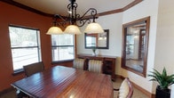 This well-appointed single-floor space exudes a warm, rustic elegance through its thoughtful use of rich wood details and carefully chosen finishes. The centerpiece of the space is a dining or conference area anchored by a substantial solid wood table surrounded by striped upholstered chairs. Above, a striking wrought iron chandelier with frosted glass pendants casts a welcoming glow, while an exposed wood beam adds architectural character to the ceiling. Bold terracotta-toned accent walls contrast beautifully with cream upper walls, creating a layered, inviting atmosphere. Multiple windows fitted with horizontal blinds line the room, allowing natural light to filter in and offering views of the surrounding exterior. A rich wood sideboard with a framed mirror above serves as an elegant focal point along one wall. Throughout the space, dark stained wood trim, baseboards, and door casings provide a consistent and polished aesthetic. A standout feature is the classic French door with a multi-pane glass grid and oil-rubbed bronze hardware, opening into a hallway finished with tile flooring and additional wood-trimmed interior windows. The corridor connects to further rooms, each framed by solid raised-panel wood doors, completing the cohesive and refined character of this inviting space.