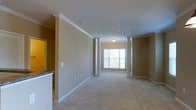 This single-floor space offers a thoughtfully designed open layout that feels both spacious and welcoming. The main living area is immediately distinguished by a set of decorative white columns that define zones within the open floor plan without compromising the sense of openness. These columns are complemented by crisp white crown molding that runs along the ceiling, adding a refined architectural touch to the warm, neutral-toned walls.

Soft, plush carpeting in a light neutral tone extends throughout the main living area, contributing to a comfortable and cohesive atmosphere. A large window at the far end of the room invites an abundance of natural light, brightening the interior and creating an airy ambiance. Recessed lighting and a flush-mount ceiling fixture provide additional illumination.

A partial view into an adjacent area reveals granite countertops, suggesting a well-appointed kitchen nearby. Wide baseboards and clean lines throughout reinforce the space's polished, classic aesthetic. The overall design strikes a balance between traditional architectural detailing and a clean, open layout, making it a functional and elegant environment suited for comfortable everyday living.