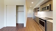 This single-floor unit presents a thoughtfully designed, open-concept layout finished with warm hardwood-style flooring throughout. The main living area is bright and airy, benefiting from a large window that frames outdoor greenery and a wall-mounted HVAC unit for year-round climate control. A sleek black-framed door provides direct access to an outdoor area, adding a welcoming sense of connectivity to the exterior.

The kitchen is neatly integrated into a dedicated alcove, offering stainless steel appliances including a refrigerator, electric range, dishwasher, and over-the-range microwave. Medium-toned wood cabinetry with open shelving complements the clean white countertops, and recessed lighting illuminates the workspace efficiently.

A sliding door reveals a generous closet with a full-width hanging rod, providing ample storage. The hallway leads to the bathroom, which features rich wood-tone cabinetry with a white countertop vanity, an inset mirror, accessibility grab bars, and the same durable hardwood-style flooring that unifies the entire space. The overall design is clean, contemporary, and highly functional.