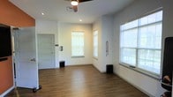 This single-floor space offers a clean, open layout with a modern, minimalist design. The interior is finished with smooth white walls and rich hardwood-style flooring that runs throughout, creating a warm yet contemporary feel. Multiple windows along the walls allow ample natural light to fill the room, enhancing the sense of openness and airiness. Recessed ceiling lights provide additional illumination, complemented by a ceiling fan that supports air circulation. A paneled door with a light finish provides access to an adjoining area or exterior, adding functional connectivity to the layout. The ceiling is standard height and well-finished, contributing to the overall neat appearance of the space. Wall-mounted fixtures and built-in ventilation details reflect thoughtful utility integration. The open floor plan offers flexibility for a variety of uses, from professional or studio purposes to general multipurpose functions. Overall, the space presents a tidy, well-maintained environment with a neutral palette that allows for easy customization to suit a range of needs.