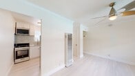 This well-appointed single-floor space offers a clean, modern aesthetic defined by light hardwood flooring, crisp white walls, and elegant crown molding that runs throughout. The open living area is bathed in natural light from a wide picture window overlooking an outdoor railing, creating an airy and inviting atmosphere. A sleek ceiling fan with a contemporary design provides both style and comfort overhead.

The kitchen is a standout feature, showcasing white shaker-style cabinetry paired with striking granite countertops and a matching full granite tile backsplash. A stainless steel undermount sink, a brushed nickel pull-down faucet, and a gas range complete this well-equipped culinary space. Natural light filters in through a window above the sink, further brightening the room.

The bedroom area is generously proportioned, featuring the same light hardwood flooring, crown molding, and a ceiling fan. A wall-mounted heating unit provides efficient climate control, and a closet with a hanging rod offers practical storage.

The bathroom is finished with classic white subway tiles surrounding a full soaking tub and shower combination, accented by brushed gold fixtures that add a warm, refined touch. Together, these spaces form a cohesive, thoughtfully updated interior with a light and neutral palette throughout.