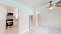 This well-appointed single-floor space offers a clean, modern aesthetic defined by light hardwood flooring, crisp white walls, and elegant crown molding that runs throughout. The open living area is bathed in natural light from a wide picture window overlooking an outdoor railing, creating an airy and inviting atmosphere. A sleek ceiling fan with a contemporary design provides both style and comfort overhead.

The kitchen is a standout feature, showcasing white shaker-style cabinetry paired with striking granite countertops and a matching full granite tile backsplash. A stainless steel undermount sink, a brushed nickel pull-down faucet, and a gas range complete this well-equipped culinary space. Natural light filters in through a window above the sink, further brightening the room.

The bedroom area is generously proportioned, featuring the same light hardwood flooring, crown molding, and a ceiling fan. A wall-mounted heating unit provides efficient climate control, and a closet with a hanging rod offers practical storage.

The bathroom is finished with classic white subway tiles surrounding a full soaking tub and shower combination, accented by brushed gold fixtures that add a warm, refined touch. Together, these spaces form a cohesive, thoughtfully updated interior with a light and neutral palette throughout.