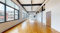 Located at 210 Pryor St SW in the heart of downtown Atlanta, this unit offers a distinctive loft-style living experience defined by its industrial architectural character. The expansive main living area is immediately striking, featuring soaring ceilings with exposed black steel beams, white-painted wood plank ceilings, and visible ductwork that celebrate the building's industrial heritage. Warm-toned hardwood floors flow throughout the open space, while a dramatic run of large multi-pane windows along one wall floods the interior with natural light and frames views of the surrounding Atlanta cityscape. A ceiling fan adds both function and charm to the airy main room. The generously sized bedroom provides ample storage with two built-in closets and maintains the same hardwood flooring found throughout. The bathroom features a modern vanity with wood-toned cabinetry, a dark countertop, an illuminated mirror, and clean tile flooring. A dedicated laundry area houses a stacked front-loading washer and dryer alongside a water heater, keeping utilities neatly contained. The overall ambiance of this loft is one of open, industrial-chic urban living, combining raw architectural details with functional, well-appointed spaces in a prime downtown location.