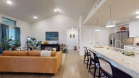 This single-floor residence showcases a well-appointed open-concept layout that seamlessly connects the living, dining, and kitchen areas. The living room stands out with soaring vaulted ceilings and large windows dressed in steel-blue drapes, flooding the space with natural light. Light wood-style flooring runs throughout the main living areas, complementing the bright, neutral color palette.

The kitchen is a functional centerpiece, featuring white shaker cabinetry, stainless steel appliances, a classic subway tile backsplash, and granite countertops. A generous island with bar seating and pendant lighting above makes it ideal for both cooking and casual dining.

The primary bedroom suite offers plush carpeting and direct access to a well-designed en-suite bathroom, which includes a dual-sink vanity with granite countertops and a glass-enclosed walk-in marble tile shower. A second bathroom provides a full soaking tub with marble tile surround. Additional rooms include a versatile office or flex space with French doors opening to the outdoors, allowing for an abundance of natural light. Secondary bedrooms are carpeted and include access to closets. The overall design balances comfort and functionality with cohesive, upscale finishes throughout.