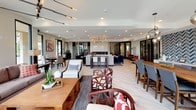 This single-story community clubhouse is a well-appointed gathering space designed to accommodate a variety of social activities. The open floor plan is expansive, with light hardwood-style flooring flowing throughout most of the space and transitioning to carpeted areas in designated lounge zones.

The main lounge area features multiple seating arrangements, including navy blue upholstered armchairs, a tan sofa, and eclectic accent chairs with bold geometric frames, all centered around wooden coffee tables. A centrally located bar with high-top seating serves as a focal point, accented by a striking blue herringbone tile backsplash and an industrial-style chandelier overhead.

A dedicated dining area toward one end of the space includes a rectangular wood dining table with tufted upholstered chairs and a large-scale abstract artwork on the wall. Black-framed glass doors and windows throughout the space invite abundant natural light and provide direct views of an outdoor pool area surrounded by palm trees. Track lighting and pendant fixtures complement the recessed ceiling lights, contributing to a warm, modern ambiance suited for both casual gatherings and organized events.