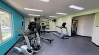 Located at 8470 Towson Blvd. in Miamisburg, Ohio, this single-floor fitness center is a well-equipped exercise facility designed to support a full range of workout routines. The space features a clean, functional layout with durable rubber flooring throughout, a textured ceiling with recessed and flush-panel fluorescent lighting, and a vibrant teal accent wall that adds a lively, energetic atmosphere.

Cardio equipment includes True-brand elliptical trainers and a treadmill, positioned to take advantage of the large windows that line the teal wall, offering pleasant views of the surrounding tree-lined grounds. A multi-station cable weight machine provides versatile strength training options, while a dedicated free weight area includes a dumbbell rack with a full set of dumbbells and a barbell with weight plates. A wall-mounted flat-screen television is available for entertainment during workouts.

Large mirrors on one wall enhance the sense of space and allow users to monitor their form. A wall-mounted fan provides additional airflow. A vending machine is conveniently accessible near the entrance. The facility is well-lit and thoughtfully organized, offering a welcoming and practical environment for both cardiovascular and strength training exercise.