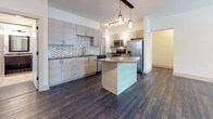 This well-appointed single-floor residence offers a thoughtfully designed open layout with consistent dark hardwood flooring that flows throughout the main living areas and bedrooms, creating a cohesive and contemporary aesthetic. The spacious living room is bright and airy, featuring a ceiling fan with light fixture, a welcoming exterior door with window, and ample wall space that allows for flexible furniture arrangements.

The kitchen is a standout feature of the home, showcasing light wood cabinetry, a striking mosaic tile backsplash, and a large granite-topped island ideal for dining or entertaining. Stainless steel appliances — including a refrigerator, range, and microwave — are complemented by pendant and track lighting overhead, adding warmth and functionality to the space. Generous counter and cabinet space make this kitchen as practical as it is attractive.

The bathroom is finished with large-format tile flooring, a granite vanity with a dark-framed cabinet, and a full soaking tub and shower combination with matte black fixtures and grab bars. Additional rooms offer clean, neutral walls, natural light through slatted blinds, and the same rich hardwood flooring. The overall ambiance is modern, clean, and move-in ready, with quality finishes throughout.