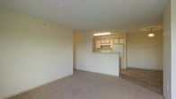 This single-floor residence offers a straightforward and functional layout designed for comfortable everyday living. The living area is finished with wall-to-wall carpet in a neutral tone, providing a clean and cohesive look that complements the light-colored walls throughout. The space feels open and uncluttered, offering a blank canvas well-suited for a variety of furnishing arrangements.

A defining feature of the layout is the pass-through kitchen, which connects to the living area via a raised counter that doubles as a breakfast bar or serving surface. The kitchen is equipped with light wood cabinetry providing ample storage, a white refrigerator, a range, and an overhead fluorescent light fixture that keeps the cooking area well-lit. The kitchen flooring transitions to tile, offering a practical, easy-to-clean surface in this high-use zone.

The textured ceiling adds a subtle architectural detail throughout, while the neutral color palette of cream and beige tones creates a warm, inviting ambiance. The overall design emphasizes practicality and livability, making efficient use of the available space. The open sightline between the living and kitchen areas enhances the sense of space and promotes a connected, sociable atmosphere within the home.