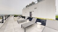 Located in Montréal, this contemporary lounge and terrace space combines industrial design with refined comfort across a generously sized single floor. The interior is defined by exposed concrete ceilings with visible ductwork and structural columns, polished concrete floors, and floor-to-ceiling windows that frame panoramic views of the surrounding cityscape. The open-plan lounge area is anchored by a striking suspended fireplace and features a distinctive circular built-in bench that wraps around a central column, offering a unique social focal point. Vibrant coral and terracotta upholstered seating, round accent rugs, and sleek side tables add warmth and color against the neutral industrial palette. A semi-private dining nook with a wooden table and bench seating is separated by glass partitions, while slatted wood screens provide subtle spatial definition throughout. The space transitions seamlessly to an expansive rooftop terrace, accessible via large sliding glass doors. The terrace is outfitted with cushioned lounge chairs, sectional seating, and planters filled with ornamental grasses and trees, all set against wide-open views of the Montréal skyline — creating an ideal environment for relaxation and social gatherings alike.