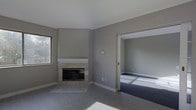 This single-story space presents a clean, understated aesthetic defined by neutral grey walls and light-toned finishes throughout. The main living area centers around a tiled fireplace with a classic white mantel surround, serving as a natural focal point. The flooring in this area transitions between tile near the hearth and smooth grey flooring extending across the room, adding subtle visual variety.

A window positioned to the left of the fireplace allows natural light to filter into the space, illuminating the room's simple, contemporary palette. The walls are finished in a consistent cool grey tone, contributing to a calm and cohesive atmosphere throughout.

An open doorway connects the main living area to an adjacent room, which features carpeted flooring in a complementary grey-blue tone. This room benefits from natural light as well, visible through what appears to be a window, and maintains the same neutral wall color as the primary space.

The overall layout emphasizes a straightforward, functional design with minimal ornamentation. The consistent use of grey tones and clean lines throughout both rooms creates a unified, modern ambiance that provides a versatile foundation for a variety of interior styles.